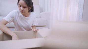 jovem mulher asiática tira coisas da caixa de papelão do pacote organizando as coisas, sente-se com o sofá atrás, sala de estar da casa nova, caixa, realocando movendo-se no conceito, fique em casa sozinho, quarto branco video