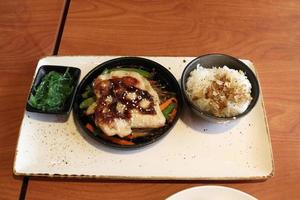 la comida japonesa puesta en un tazón negro está en un plato blanco e incluye arroz blanco y cubierto con pescado seco japonés, pollo terriyaki a la parrilla y verduras asadas debajo. foto