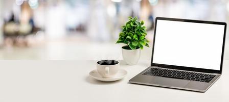 portátil de pantalla blanca vacía, taza de café y jarrón colocados en un escritorio blanco en la oficina. el concepto de negocio, tecnología, internet, diseño, programador. primer plano, enfoque selectivo, fondo borroso foto