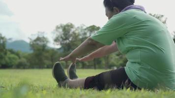 Asian active overweight senior female stretching her legs hand to toe sit on the ground, grass field trees and sky after exercise at the outdoor park, old age life relaxation after retired, side view video