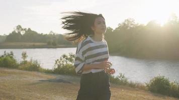 menina asiática correndo ao ar livre está ao longo do córrego do rio sente a brisa do vento ao redor, atividade ativa ao ar livre, com bela luz do sol no fundo vitalidade saudável, vibração feliz, câmera lenta video