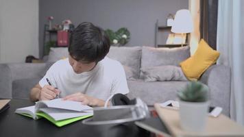 junge glückliche asiatische Studentin macht gerne Selbststudium zu Hause im Wohnzimmer, verbessert Kenntnisse und Fähigkeiten, versteht sich im Unterricht mit dem Schreiben einer kurzen Notiz zum Lehrbuch, komfortables, gemütliches Zuhause,