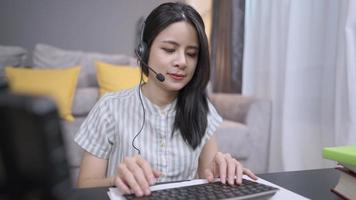 Asian woman wear headset with microphone talking interact on live call with customer, working at home, typing on  wireless keyboard, Online freelancer answering question, register operator service video