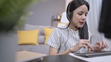 Asian young female wear headset talking on conference call, microphone voice interact with client, typing on the keyboard, Online customer care live chat with staff, stay working from home living room
