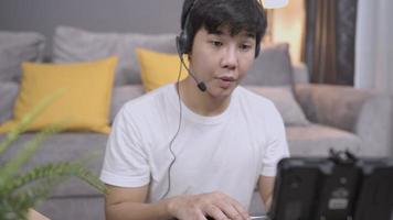 un joven asiático feliz usa auriculares que se comunican por llamada de conferencia en la oficina de su casa, videochat de servicio al cliente del operador o clase de curso de idioma a distancia con un profesor en línea, trabajo a tiempo parcial