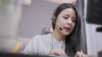 asiatische junge Dame trägt ein Headset, spricht und tippt die Tastatur ein, unterstützt das Voice-Callcenter, Interaktion mit dem Ausbilder, Online-Tutorenkurs, Live-Chat-Diskussion mit Mitarbeitern, Beratung von zu Hause aus video