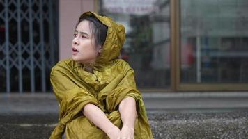 young asian female in yellow hoody rain coat sit down waiting on the street side on the rainy day, stuck in the rain, self protection from, wet clothes stuck in the rain, wait for rain to stop video