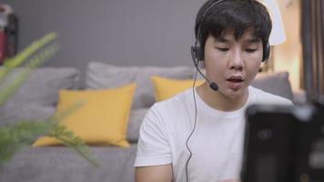 un jeune homme asiatique porte un casque d'écoute ayant une conversation avec des clients, une interaction d'appel vocal avec un microphone avec le client, un service client en ligne, un chat en direct avec le personnel, du travail à domicile, des vêtements décontractés à la maison video
