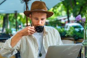 joven inconformista con estilo en el trabajo del sombrero con la computadora portátil al aire libre en el café de la calle foto