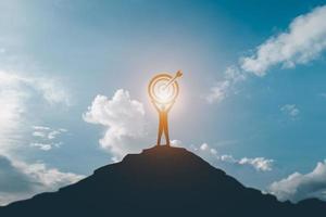 la silueta de un hombre de negocios que sostiene un tablero de destino en una montaña de azul y luz solar es un símbolo de éxito en el liderazgo con un propósito. foto