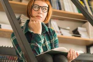Young redhead woman in glasses read book in the library photo
