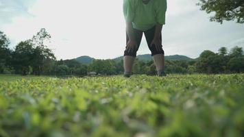 An oversized female in casual clothing stretching a leg muscles after workout on green fresh meadow in natural park, hands touches on knees with bending body forward, relaxing exercise, health care video