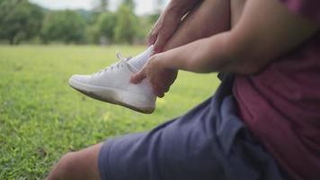 Asian man having ankle injury during jogging exercise at the park sit down on tree trunk green grass on the background, ankle pain, joint ligament problem, unexpected accident, legs muscles tendon video