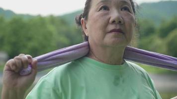 primer plano de una anciana asiática haciendo ejercicio en el parque, una señora madura estirándose con una toalla, actividad al aire libre relajándose después de hacer ejercicio, jubilada activa optimista, longevidad de bienestar de la salud video