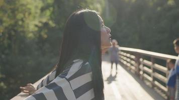 Young asian woman feeling relax enjoying morning sunlight inside park creek, standing on wooden bridge, people walking and relaxing on the background, enjoy herself on the weekend outdoor activity video