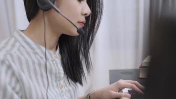 vista laterale donna asiatica operatore di call center lavoratore, primo piano auricolare femmina che comunica tramite teleconferenza con il cliente, lavoratore a domicilio, colloquio di lavoro o corsi online a distanza, risolvere il problema dei clienti video