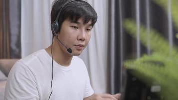 Asian young man wear headset doing work from home call operator, microphone voice call interaction with customer, live chat with staff, late night shift, casual  clothing doing working at home