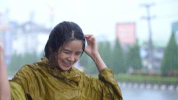 retrato de uma mulher asiática alegre na capa de chuva amarela molhada pulando na chuva forte, se divertindo se molhar no dia chuvoso, dança sorridente engraçada, braços estendidos, trabalhador alivia o estresse emocional video