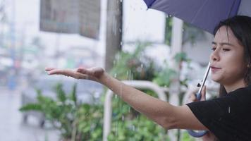 joven asiática sosteniendo paraguas sentada esperando en el lado de la calle con lluvia torrencial, temporada de tormentas lluviosas, extiende su mano tocando gotas de lluvia, atrapada en la lluvia, cara de curiosidades positivas