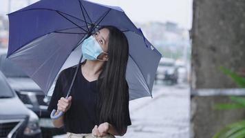 una joven asiática usa una mascarilla protectora que sostiene un paraguas azul parado en la calle, se baña en la temporada de lluvias, llueve a cántaros, corre el riesgo de enfermarse, llueve fuerte, extiende la mano con curiosidad video