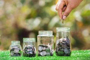 hand holding coins putting into jug glass with money stack on green grass. concept saving finance and accounting concept photo