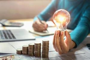 business woman hand holding lightbulb with coins stack on desk. concept saving energy and money photo