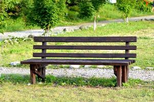 banco de madera marrón en el parque. día soleado de verano. hierba verde y árboles. zona de descanso y relax. banco vacío para sentarse. material exterior de madera. foto