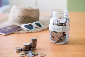 Selective focus at jar glass. Glass full of money coin with blurred straw hat, sunglasses and passport mock up at the background. Money saving plan for holiday or vacation concept. photo