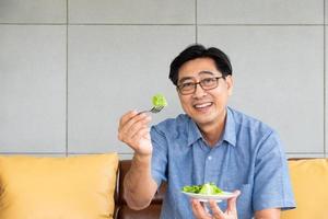 Senior Asian men sitting on the sofa at home while eating green vegetable salad and organic orange juice for breakfast. Healthy eating lifestyle for Elder people concept.