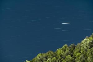 larga exposición del cielo nocturno con las estrellas moviéndose como resultado de la rotación de la tierra con una montaña forestal en primer plano foto