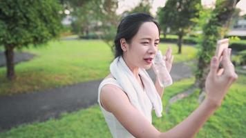 jovem mulher asiática fazendo videochamadas com amigos durante o exercício no parque, segurando uma garrafa de água de plástico, aproveite o exercício compartilhando experiências de vida saudáveis, conversando on-line, emoção positiva