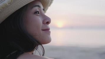 Close up head shot of young asian female enjoy sit down on sunset beach, wearing summer straw beach hat, traveling on summer vacation, smiling face outdoor nature relaxation happiness, freedom life