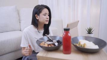Young asian woman eating healthy food alone sit down at apartment living room, budget single life,  with couch behind showing simple life, watching online content via tablet, food delivery service video