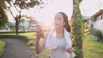 joven mujer asiática activa haciendo videollamadas con amigos mientras hace ejercicio en el parque con la puesta de sol de fondo, compartiendo historias de estilo de vida saludable, tecnología humana moderna, adaptación al nuevo distanciamiento normal