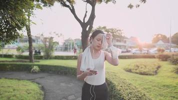 jonge aziatische vrouw in sportkleding wandelen in het park tijdens zonsondergang gouden uur, ontspannende wandeling zweet afvegen na hard werk oefeningen, met behulp van telefoon zingen op de muziek van oortelefoons video