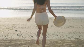 una vista trasera siguiendo a una chica sexy en bikini sosteniendo un sombrero y corriendo alegremente hacia el agua del mar, caminando contra la luz del sol, hermosas y calientes vacaciones de verano, un cabello largo y negro que fluye en el aire