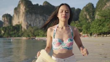 una chica esbelta del este de Asia con un lindo bikini y un pantalón corto blanco corriendo lentamente en la costa de una playa, una mujer alegre y segura de sí misma girando completamente su cuerpo, modelando pasarela, recursos tropicales naturales video