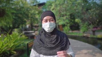 slow motion-porträtt av ung asiatisk muslimsk kvinna lägre ner ansiktsmask och leende som går i parken en solig dag, ny normal coronavirus covid-19-pandemi, förebyggande av infektionssjukdomar, lyckligt liv video