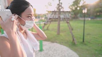 ung asiatisk kvinna sätter på sig en skyddande ansiktsmask efter att ha tränat, solnedgångsljus på bakgrunden, håller mobiltelefonen, förbli frisk under pandemin covid19 coronavirus, social distansering, sidovy video