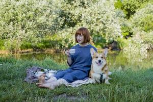 mujer joven vestida de retro con un gracioso perro corgi en el picnic, una mujer con un lindo perro bebe té inglés en el parque foto