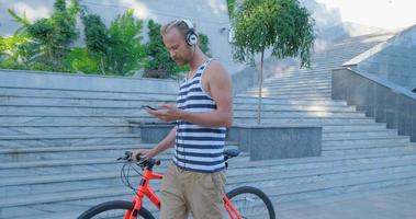 Young handsome male with colorful bicycle and headphones listen to music and ride by the summer streets