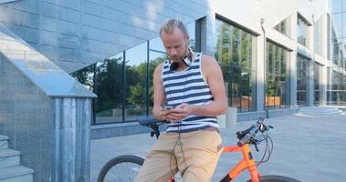 junger hübscher mann mit buntem fahrrad und kopfhörern hört musik und fährt durch die sommerstraßen video
