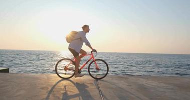 Young handsome male in casual wear ride on the colorful bicycle on the morning beach against beautiful sunset and the sea