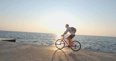 joven apuesto hombre en ropa casual paseo en la colorida bicicleta en la playa de la mañana contra la hermosa puesta de sol y el mar video