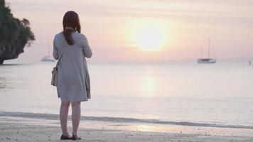 Young asian woman enjoying moment during sunset at the island beach, using smartphone taking pictures of beautiful ocean scenic, tropical island travel destination, single woman lone journey video