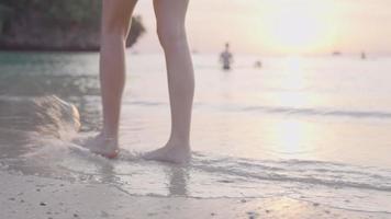 kvinnliga ben barfota promenader längs stranden med plaskande vågor, vacker solnedgång gyllene solljus reflekterar havsvatten, sommar tropisk ö strand, avkopplande promenad, närbild från sidan underkroppsdel video