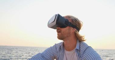 Porträt eines jungen gutaussehenden Mannes am Strand mit Virtual-Reality-Brille, schöner Sonnenaufgang im Hintergrund