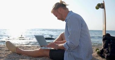 Young handsome male on the summer beach work with laptop during sunrise video