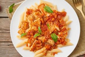 Penne pasta, chicken or turkey fillet, tomato sauce with basil leaves on old rustic wooden background. Top view, close up