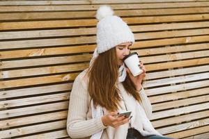 Beautiful young girl drinking coffee, tea from plastic mug in autumn, winter and talking on mobile phone. Woman with long hair sitting on bench in autumn or winter, basking in hot drink, copy space